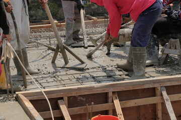 construction workers in boots using a large pipe and pump to pour wet, fluid concrete onto a floor slab at a building site