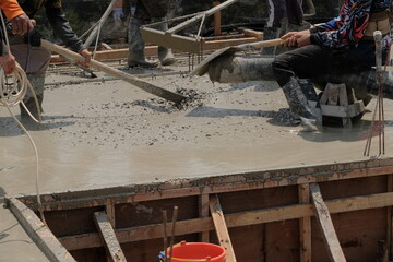 construction workers in boots using a large pipe and pump to pour wet, fluid concrete onto a floor slab at a building site