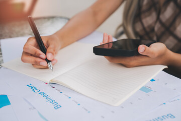 Person writing in notebook with pen and holding smartphone, business chart and documents on table, close up of hands working on financial planning and analysis