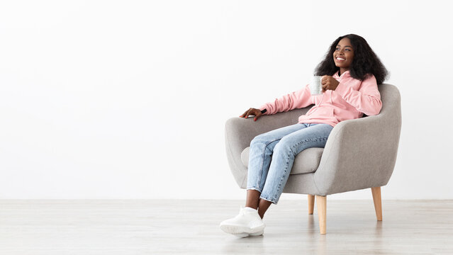 A young woman sits comfortably in a soft gray chair, smiling as she enjoys a cup of her drink in a bright, minimalistic room. Her casual attire adds to the relaxed atmosphere. - Powered by Adobe