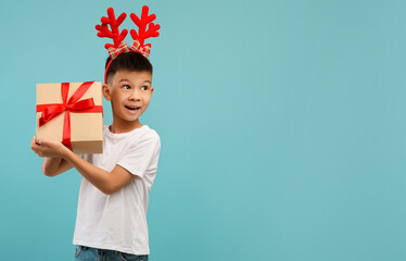 Fototapeta premium A cheerful child wearing festive reindeer antlers is carrying a wrapped gift box. The bright blue background creates a joyful holiday atmosphere.