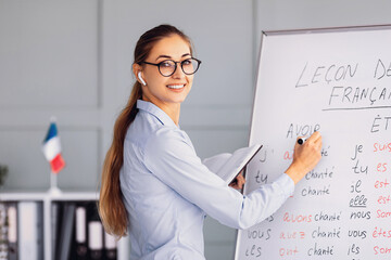 A teacher is demonstrating a French lesson on a whiteboard, writing down verbs. She is engaged and smiling, using a tablet for notes. The classroom has educational decor and a French flag in view.