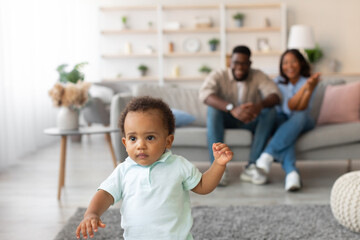 A joyful black toddler takes his first steps in the living room. His parents cheer him on from a distance, celebrating this milestone moment at home filled with love and support.