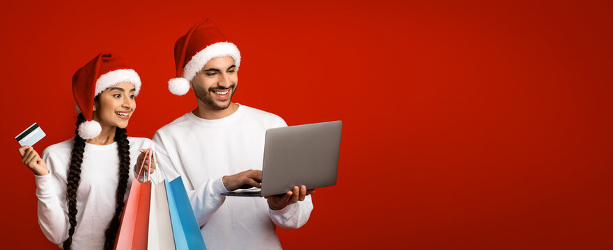 A cheerful couple wears Santa hats while shopping online. They hold shopping bags and a credit card in a vibrant red setting, capturing the spirit of Christmas shopping.