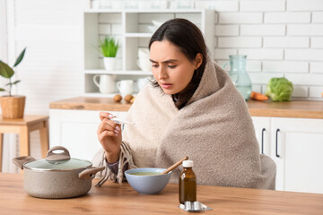 Sick young woman with thermometer and soup at table in kitchen