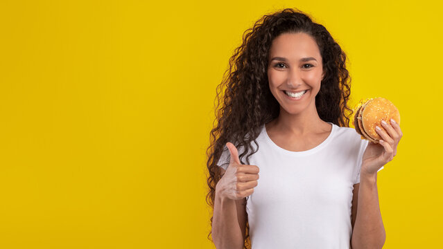 A cheerful young woman with curly hair is smiling and holding a hamburger. She is showing a thumbs up with her other hand, creating a fun and positive vibe. - Powered by Adobe