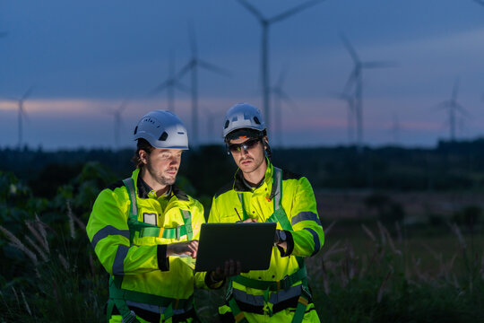 Renewable energy engineers work together on a wind farm at dusk, computerizing data and performance of the power generation system to support sustainable clean energy development.