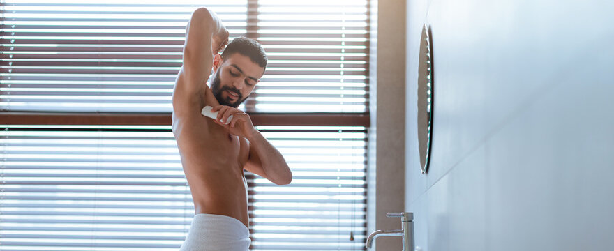 A young man is applying deodorant in a stylish bathroom. Sunlight streams in through the window blinds, highlighting his grooming routine. His expression is focused as he prepares for the day.