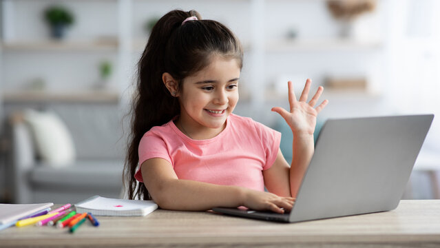 A cheerful girl with long dark hair sits at a wooden table, eagerly waving to someone on her laptop. Colorful markers are scattered nearby, reflecting her playful mood.