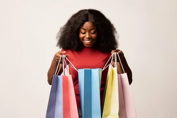Fotobehang Muziek Joyful African American woman holds colorful shopping bags with excitement, looking inside with delight. This scene captures the thrill of seasonal sales in a vibrant setting.  © Prostock-studio
