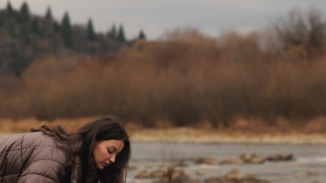 Woman setting up a camping tent by the river during an autumn outdoor adventure, wearing a warm puffer jacket while preparing her campsite in a peaceful natural landscape before solo hiking