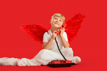 Obraz premium Cute little happy boy dressed as cupid with retro phone sitting on red background. Valentine's Day celebration