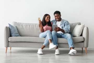 Portrait of smiling black guy and lady shopping together on Internet, using digital tablet and credit card, sitting on sofa in living room, looking at screen and smiling, buy gifts, free copy space