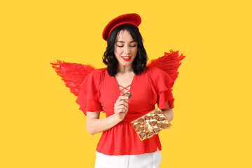 Obraz premium Young happy woman dressed as cupid with heart-shaped chocolate candies on yellow background. Valentine's Day celebration