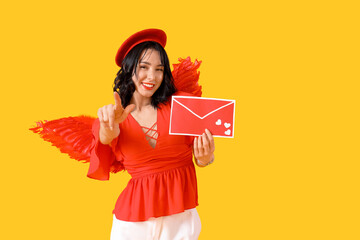 Obraz premium Beautiful young happy woman dressed as cupid with love letter pointing at viewer on yellow background. Valentine's Day celebration