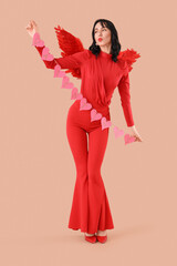 Obraz premium Beautiful young happy woman dressed as cupid with heart-shaped balloons blowing kiss on beige background. Valentine's Day celebration