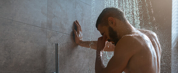 A man stands under warm water in a modern bathroom, using the shower to relax after a long day. Soft light creates a tranquil atmosphere as he enjoys this calming moment. © Prostock-studio