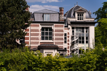 The historic facade of a brick and stone building with contrasting horizontal white bands, framed by bright green foliage