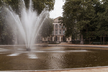 A large, splashing fountain centered in a pond in a leafy urban park with a classical building visible in the background
