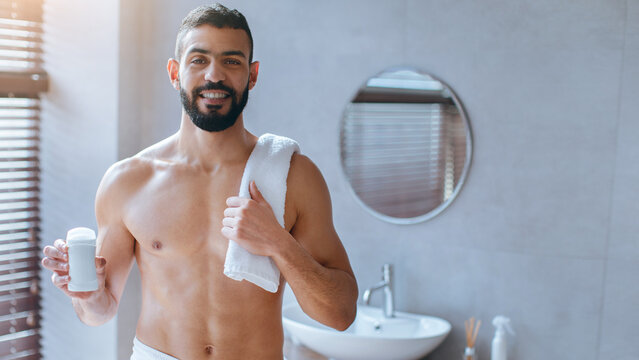 A man stands in a modern bathroom, smiling while holding a skincare product. He has a towel draped over one shoulder and appears relaxed, indicating his morning routine and self-care focus.