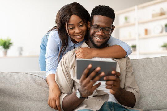 Young black couple relaxes on a sofa in a cozy living room, with the woman smiling while embracing the man who is focused on the tablet. They are having fun together.