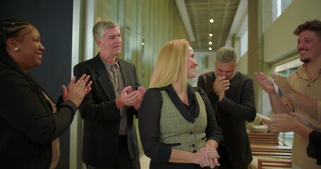 Smiling female professional receiving applause from coworkers in office hallway, diverse business team showing appreciation and support in modern workplace