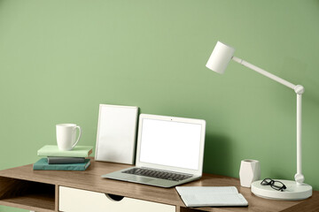 Modern workplace with laptop, lamp and books near green wall in room
