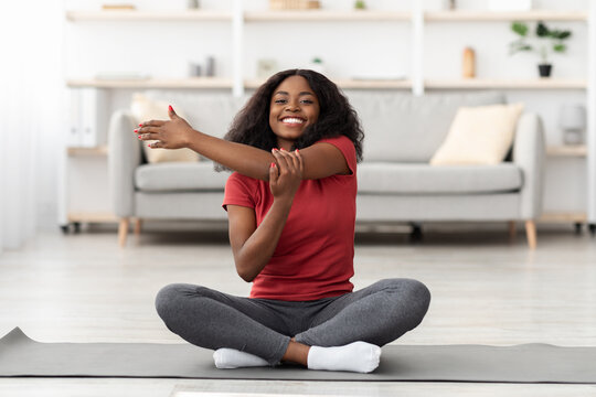 A young woman sitting cross-legged on a yoga mat stretches her arm with a smile. She appears relaxed in a bright, inviting room with modern decor.