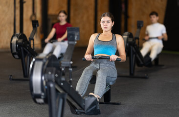 Woman performs exercises on a rowing machine, keeping the pace. Athletic woman in crossfit training doing intense exercises. Client in the gym monitors the figure and goes to training