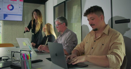 Smiling male team member types on laptop during collaborative office meeting while colleagues engage with data presentation on screen