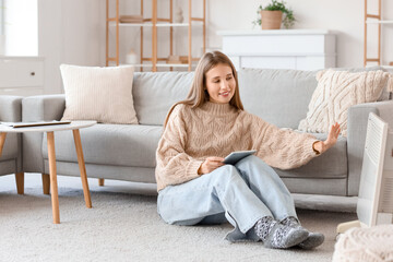 Obraz premium Young woman with tablet computer warming near electric convector heater at home