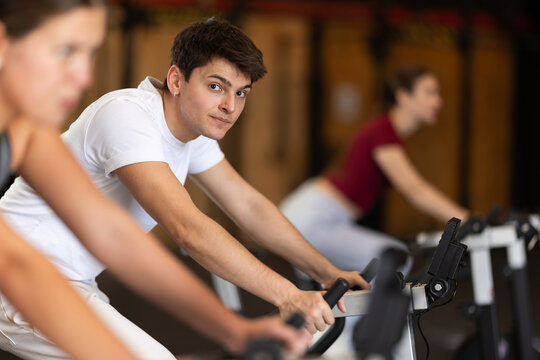 Guy hardly performs exercises on stationary bicycle during fitness class. Gym and fitness equipment, visitors are engaged in physical exercises during training