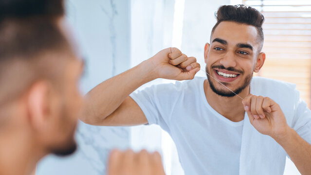 A young man smiles while flossing his teeth in a bright bathroom. The natural light enhances the cheerful atmosphere, promoting good dental care habits. - Powered by Adobe
