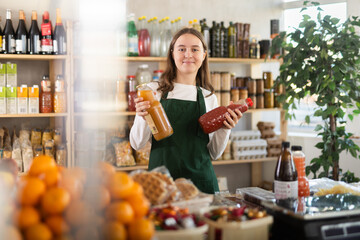 Teenager girl seller stands in trading hall of store with goods in hand, wearied in apron stands in trading hall of minimarket, ready to greets visitors and offer wine with sweet snacks in selling