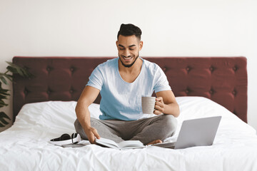 A man sits comfortably on a bed, smiling as he reads a book while holding a cup of coffee. A laptop is open beside him, creating a relaxed morning atmosphere.