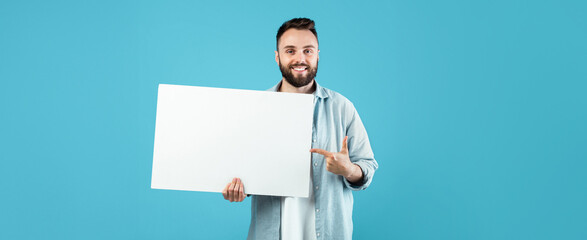A young man stands against a bright blue background, happily pointing at a blank white board he is holding. He looks friendly and ready to share a message or information.