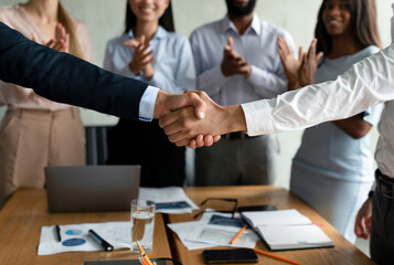Fototapeta premium Two men shake hands in a modern office after finalizing an agreement. In the background, diverse team members applaud in celebration of their successful collaboration.