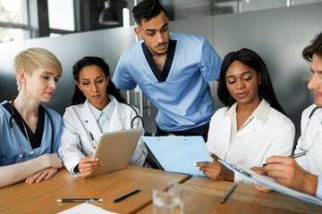 Professional international team of doctors men and women in uniforms sitting at table, using digital tablet, taking notes at medical charts, having morning breefing, clinic interior, closeup