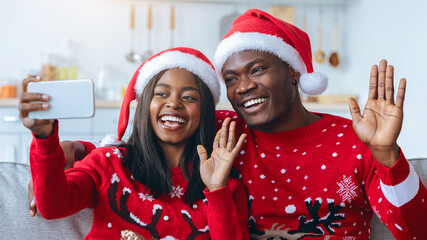 A joyful couple sits on a couch, dressed in Christmas sweaters and red Santa hats. They are smiling widely and waving at the camera while taking a selfie. The room is warmly decorated for the holiday.
