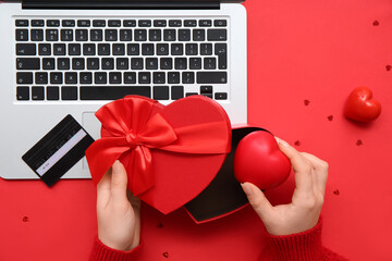 Female hands with gift box, heart, credit card and modern laptop on red background, closeup....