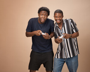Two young Black men stand closely together in a studio, smiling and holding video game controllers as they play. © The Yudel Media