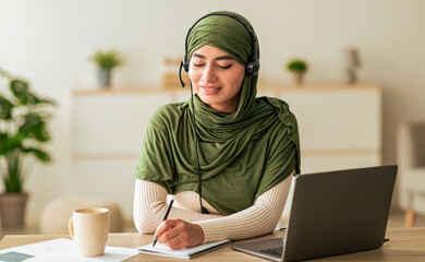 A young woman wearing a green headscarf sits at a desk. She is focused on her work, using a laptop and writing in a notebook. A cup of coffee is nearby, and plants decorate the background.