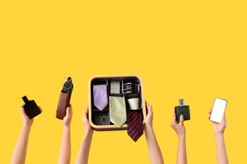 Young women holding bottles of perfume, case with eyeglasses, mobile phone and box with men's ties on yellow background
