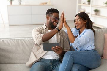 An excited African American couple sits on their couch, high-fiving each other in celebration. They are using a tablet, clearly enjoying their success together in a bright and inviting living room.