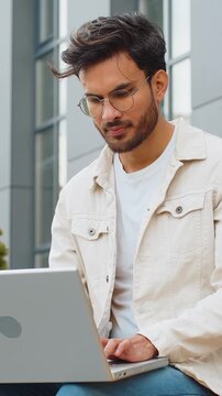 Happy young Indian man freelancer working on laptop computer sends messages, makes online purchases, watching movies, reading email outdoors. Arabian Hindu guy sitting on street bench. Business people
