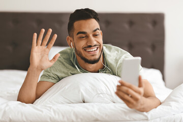 A young man is lying on his bed with earbuds in, smiling and waving at his phone during a video call. Natural light fills the room, creating a cozy atmosphere.