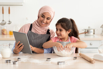 A joyful mother and daughter are baking together in a warm kitchen. The mother is holding a tablet, guiding the daughter as she mixes dough. Both are smiling and having fun.