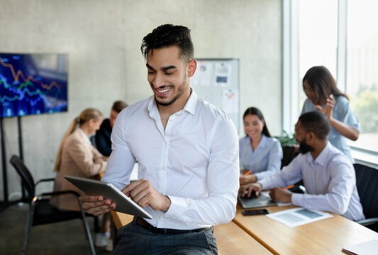 In a bright office, a young Arab man smiles while using a digital tablet. He interacts with colleagues engaged in a corporate meeting, showcasing teamwork and modern technology.