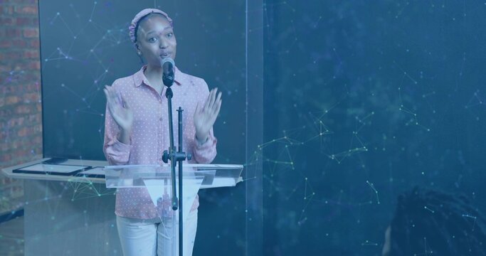 Speaking woman gesturing on stage with clear lectern and microphone, wearing pink polka-dot shirt