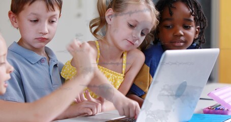 Gathering children around silver laptop at school table, girl in yellow polka top touching keyboard